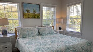 A bedroom with a large bed, two bedside tables with lamps, a painting on the wall, and windows with blinds. - The Folly Jewel -  Sleeps 6 (Folly Beach)