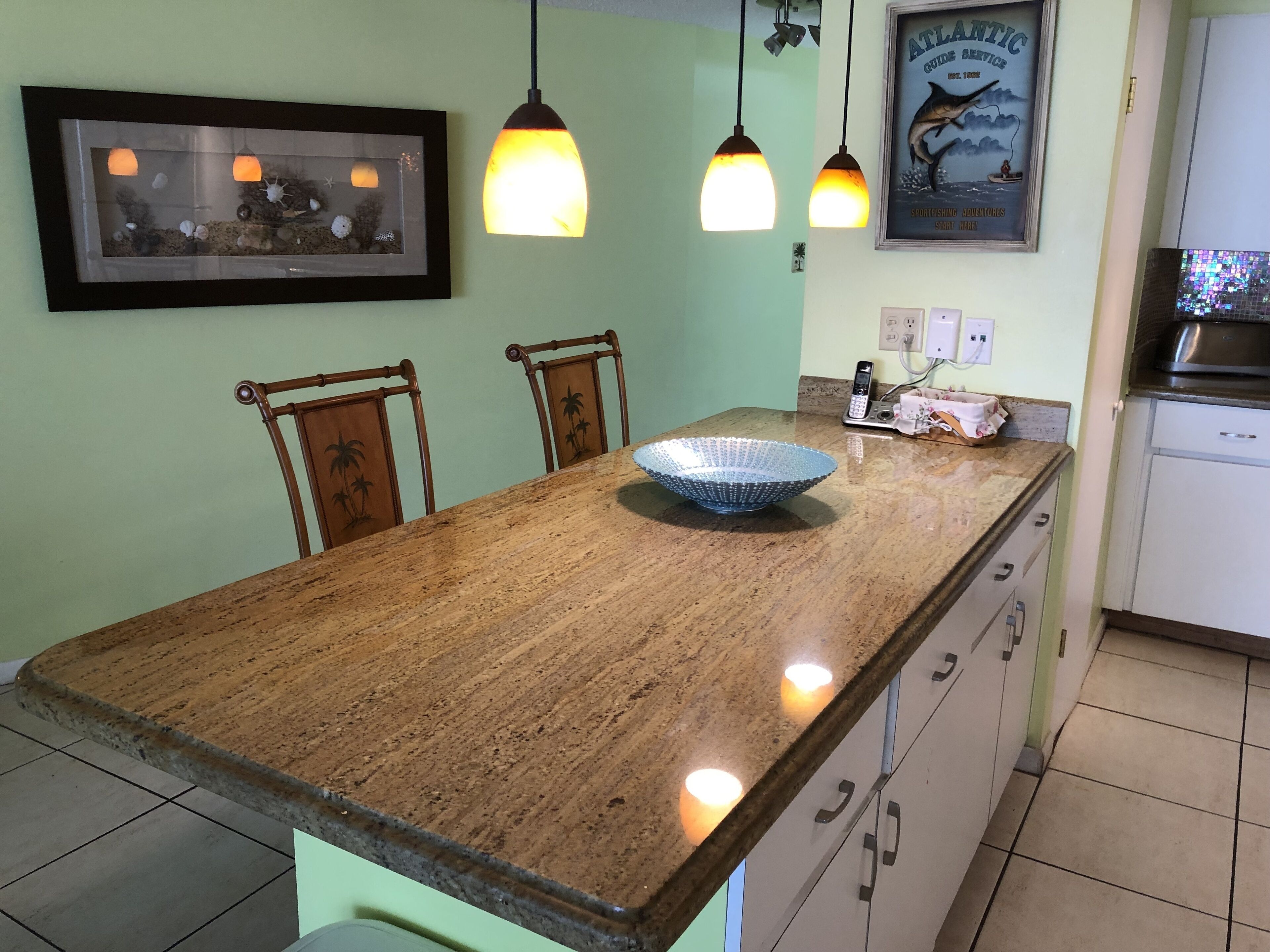Une cuisine avec un plan de travail en granit, des chaises en bois, des luminaires suspendus, un tableau encadré, une affiche et un évier.