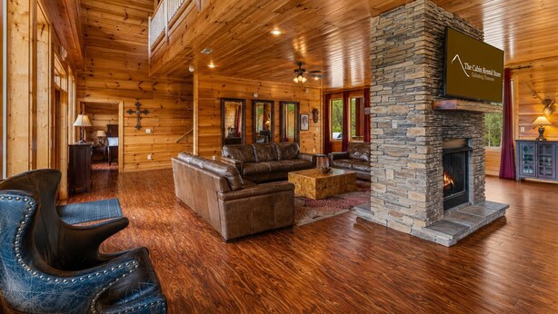 A cabin interior with wooden walls, a stone fireplace, leather furniture, and a balcony. - Foxwood Mansion | Sleeps 44 | Amazing location (Gatlinburg)