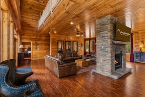 A cabin interior with wooden walls, a stone fireplace, leather furniture, and a balcony.