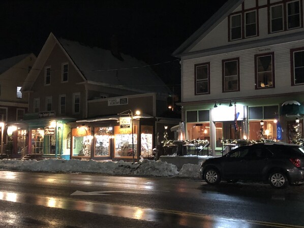 A street view at night with buildings, including a pharmacy and a restaurant, and a parked car.