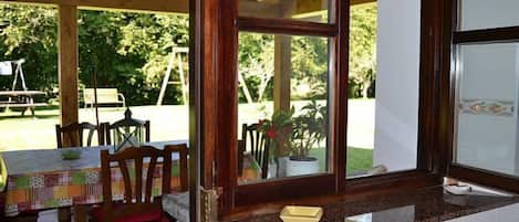 An indoor dining area with wooden chairs and a table, a window with a view of greenery, and a kitchen counter with a sink.