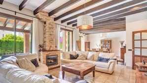 A spacious living room with a stone fireplace, large windows, and a high ceiling with exposed beams.
