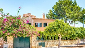 A Mediterranean-style house with a stone facade, a tiled roof, and a garden with flowering plants and trees.