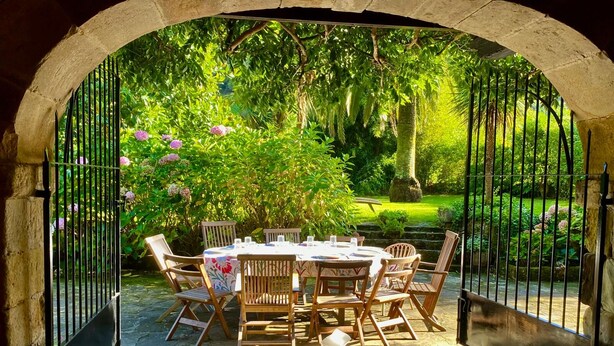 An outdoor dining area with a stone table and wooden chairs, surrounded by lush greenery and a stone archway. - Llanos De Penagos: Mansion - THE BUMBARO'S HOUSE (Llanos de Penagos)