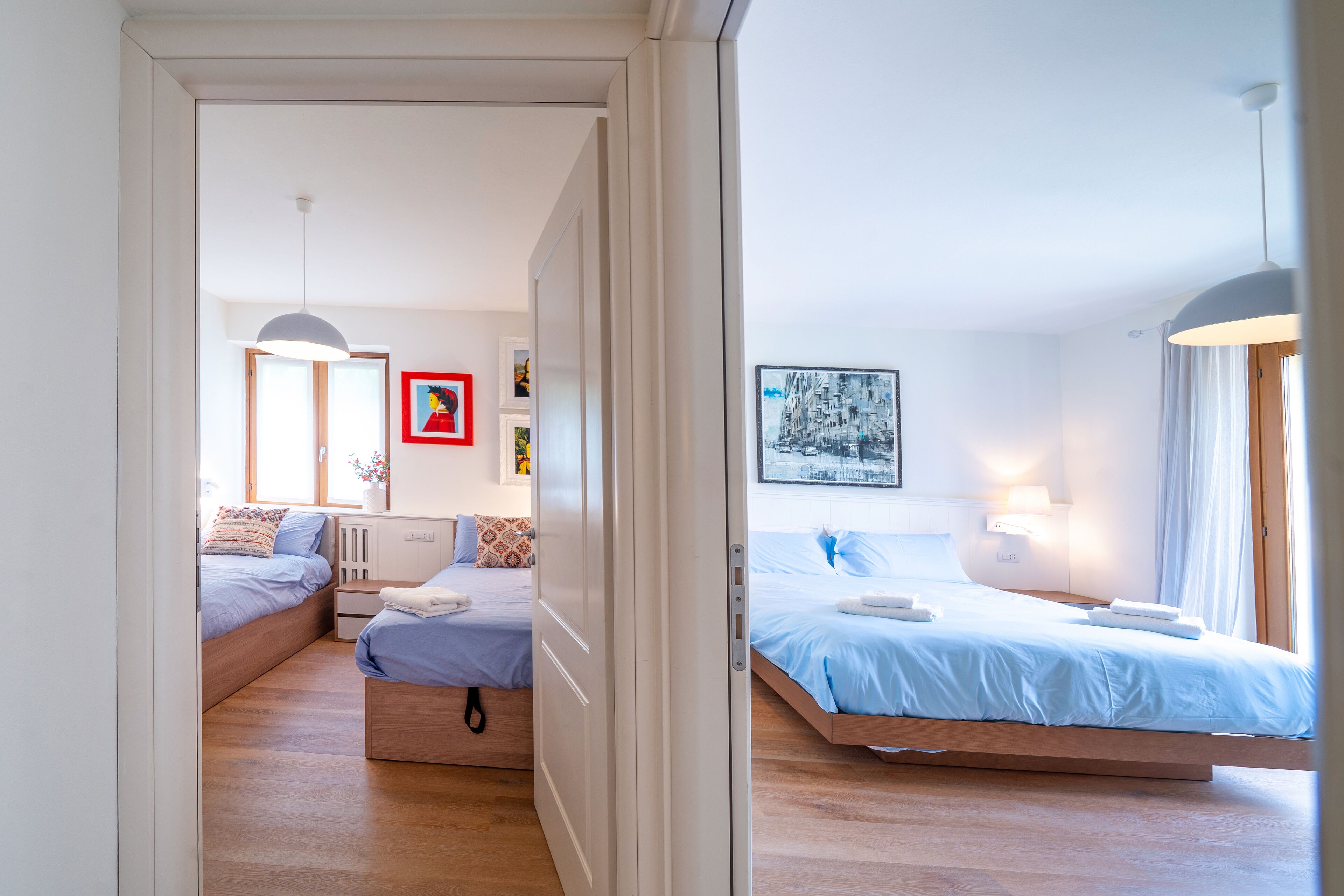 Une chambre moderne avec un grand lit, un lit plus petit et un canapé-lit. On y trouve deux tables de chevet avec des lampes, une fenêtre avec des rideaux et des œuvres d’art accrochées aux murs.