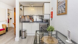 Una cocina compacta con armarios blancos, electrodomésticos de acero inoxidable y un pequeño comedor con mesa de vidrio y cesta de mimbre.