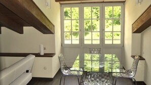 A dining area with a glass table and chairs, wooden beams, and a view of greenery through a large window. - Rural apartment Casona la Sierra for 5 people (Cudillero)
