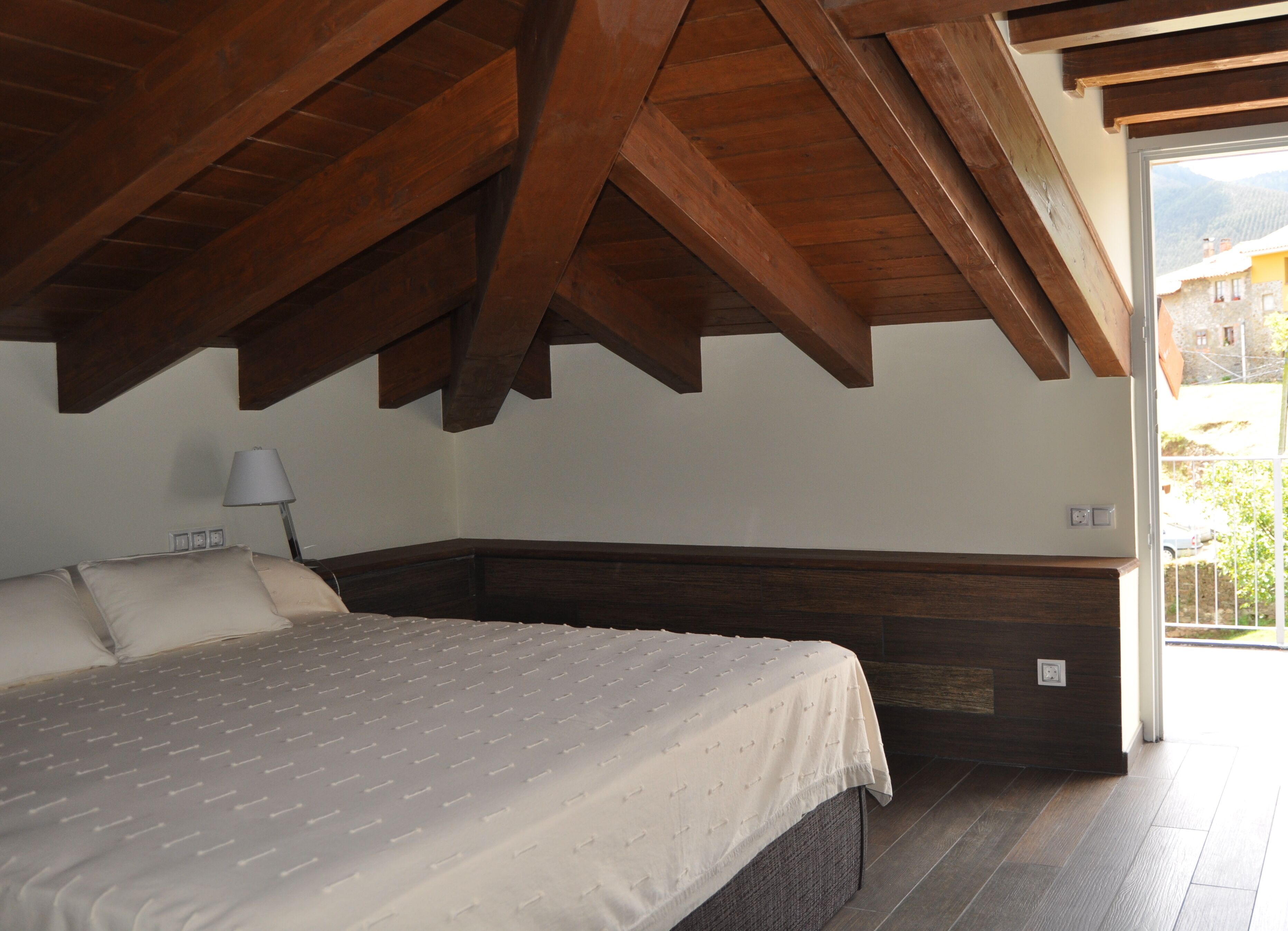 Een slaapkamer met een bed, houten balken in het plafond, een nachtkastje en een raam met uitzicht op gebouwen en groen.