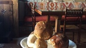 Un mur de pierres apparentes, une table en bois recouverte d’une nappe à motifs et un présentoir à gâteaux blanc sur lequel est disposé du pain.