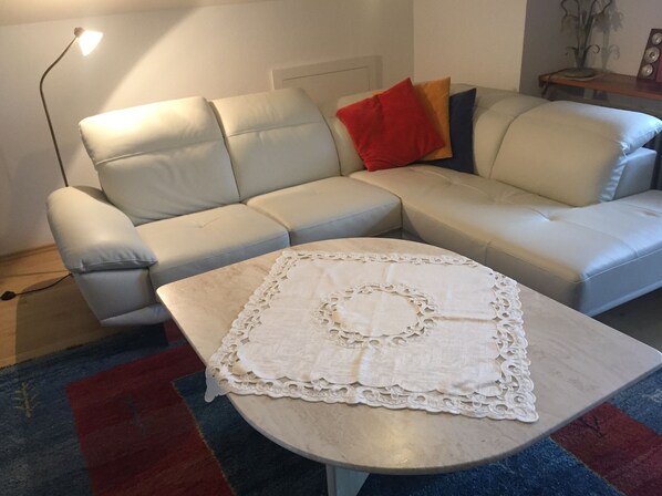 Un salon avec un canapé beige, une table basse blanche ornée d’un chemin de table en dentelle, et un tapis aux motifs rouges et bleus.