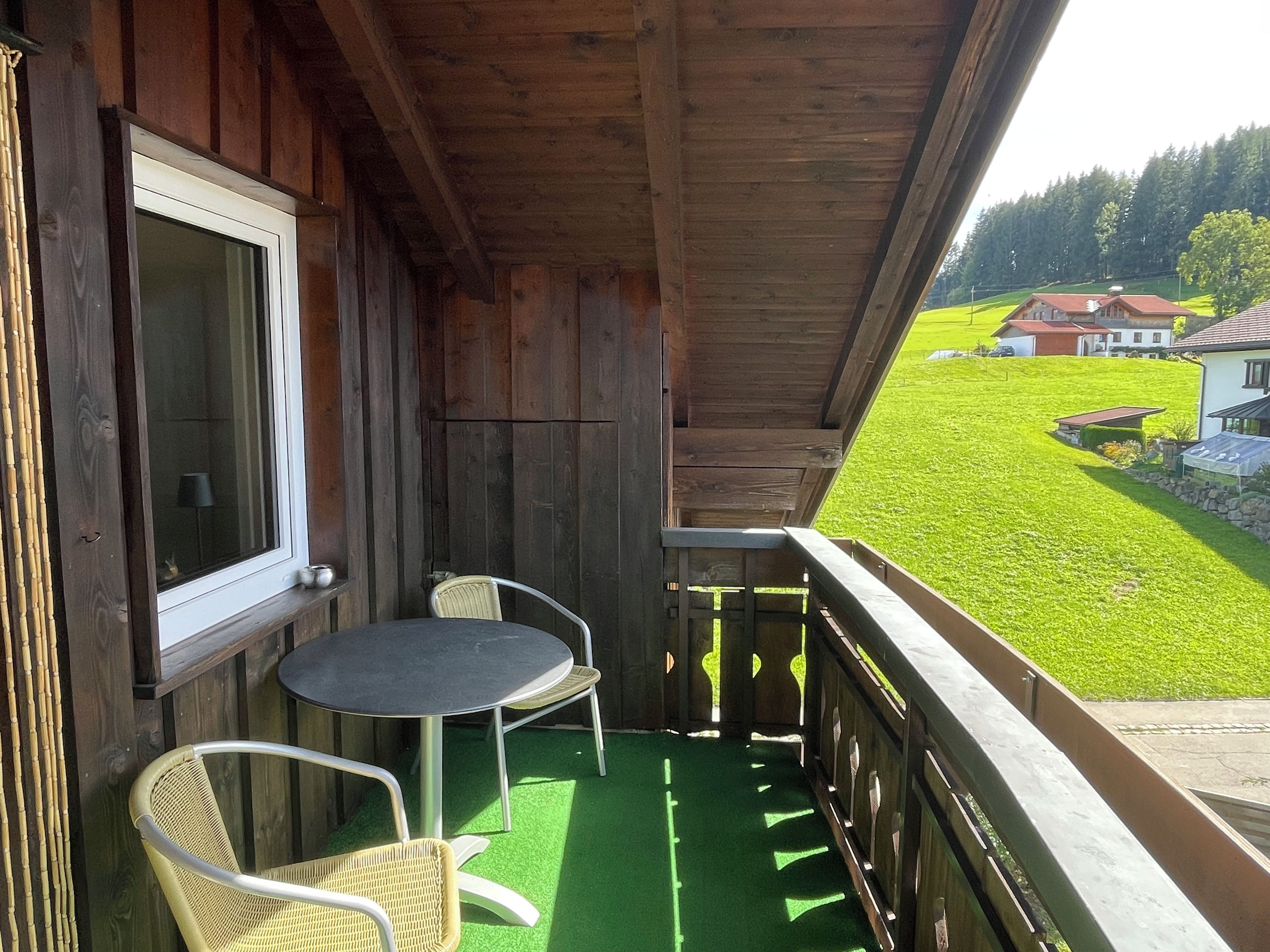 A wooden balcony with a table and two chairs, overlooking a green field and houses.