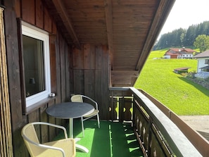 A wooden balcony with a table and two chairs, overlooking a green field and houses. - Beautiful 65 sqm apartment (Ofterschwang)