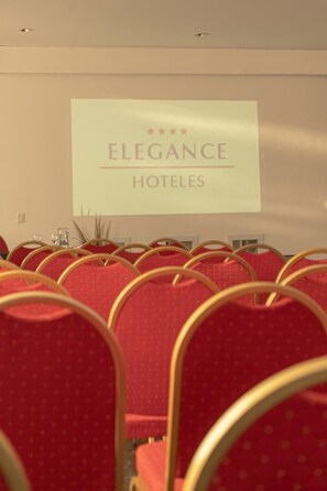 Rows of red chairs with gold frames, a projection screen displaying 'Elegance Hotels' text, and a plain wall.