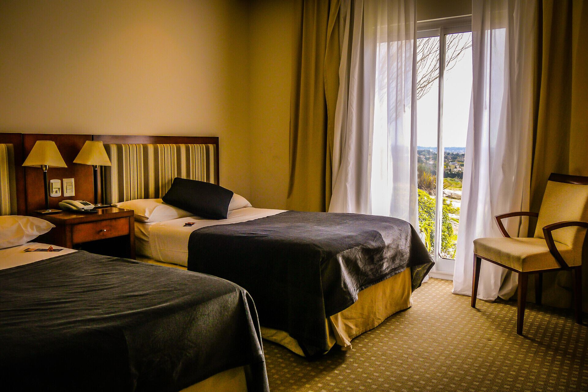 Habitación de hotel con dos camas, una silla, una mesita de noche con lámpara y vistas al exterior por la ventana.