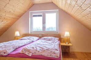 Een gezellig slaapkamer onder het dak met een schuin houten plafond, een groot raam met uitzicht op bomen, een bed met bloemenbeddels en twee bedlampen.