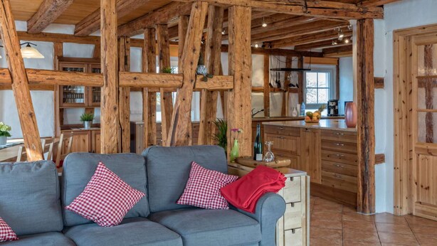 A rustic interior with exposed wooden beams, a grey sofa with checkered pillows, and a kitchen area with wooden cabinets and a window. - Exclusive Country house Seewald - quiete area, Whirlpool, Sauna, large garten, bbq, wifi (Seewald)