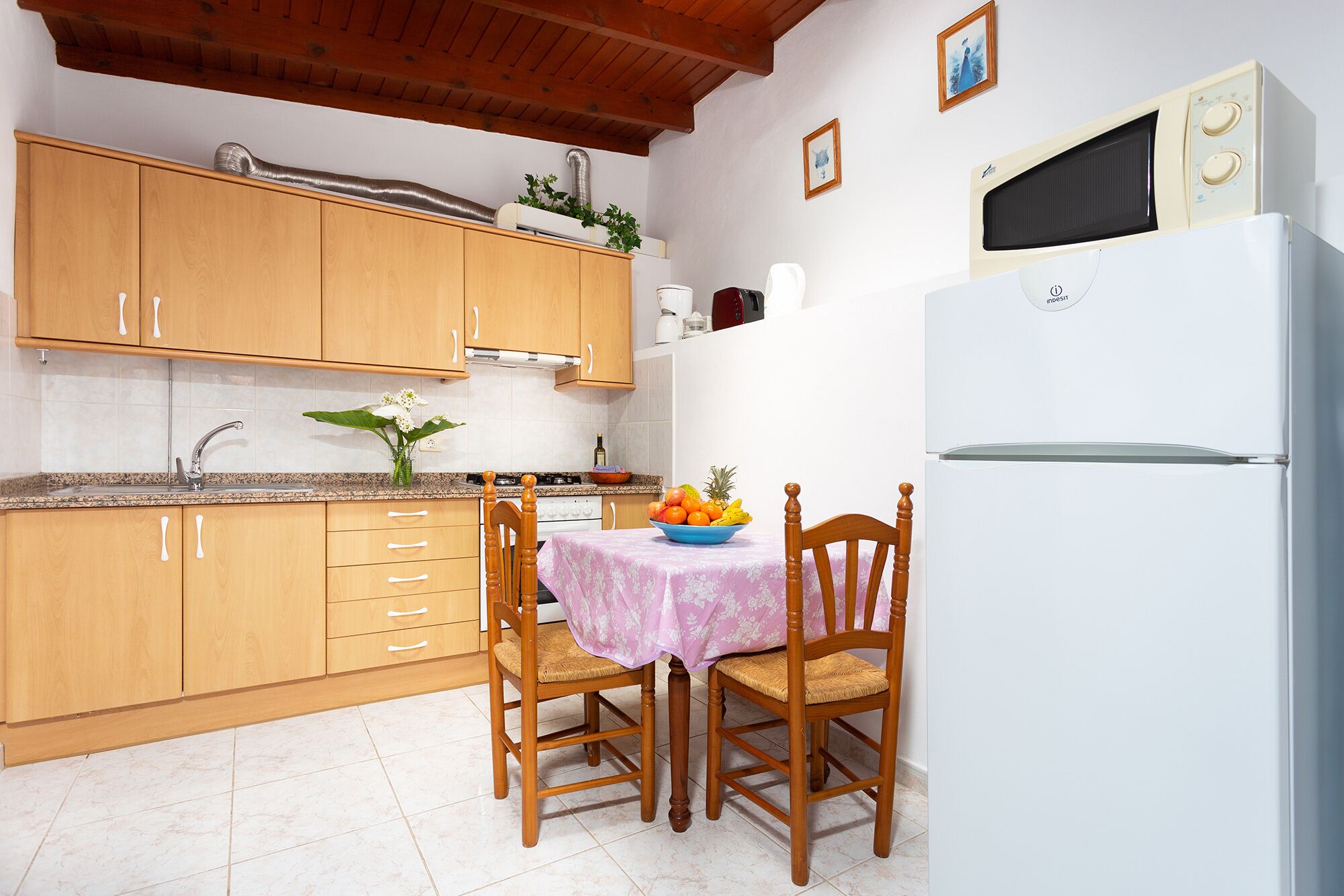 Een keuken met houten kastjes, een witte koelkast, een eettafel met stoelen en een magnetron boven het aanrecht.