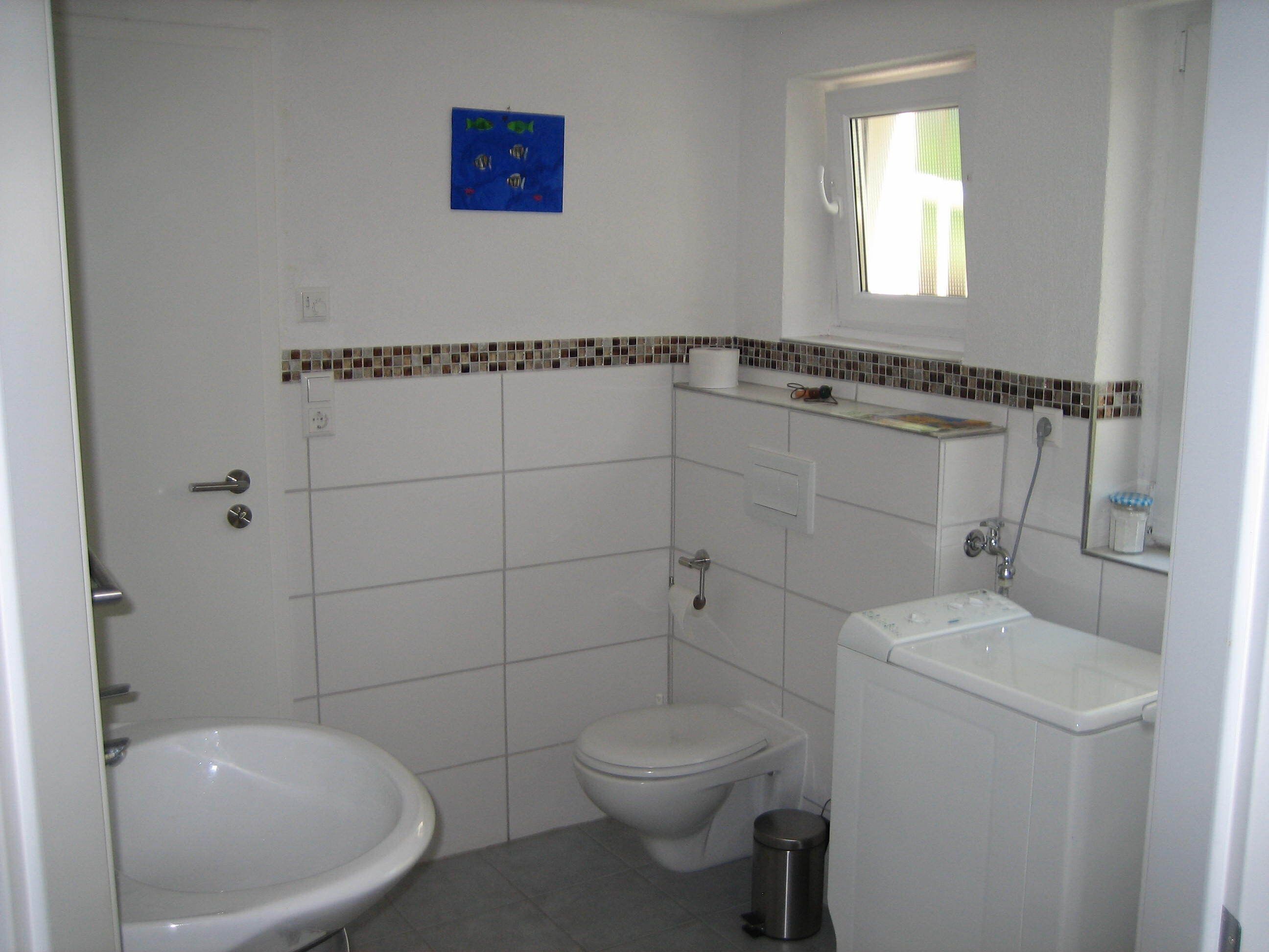 A small, white bathroom with a toilet, bidet, and sink. There is a small window above the sink and a blue sign on the wall.
