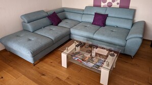 Ein graues Ecksofa mit zwei violetten Kissen, ein Glastisch mit Zeitschriften und ein Holzboden.