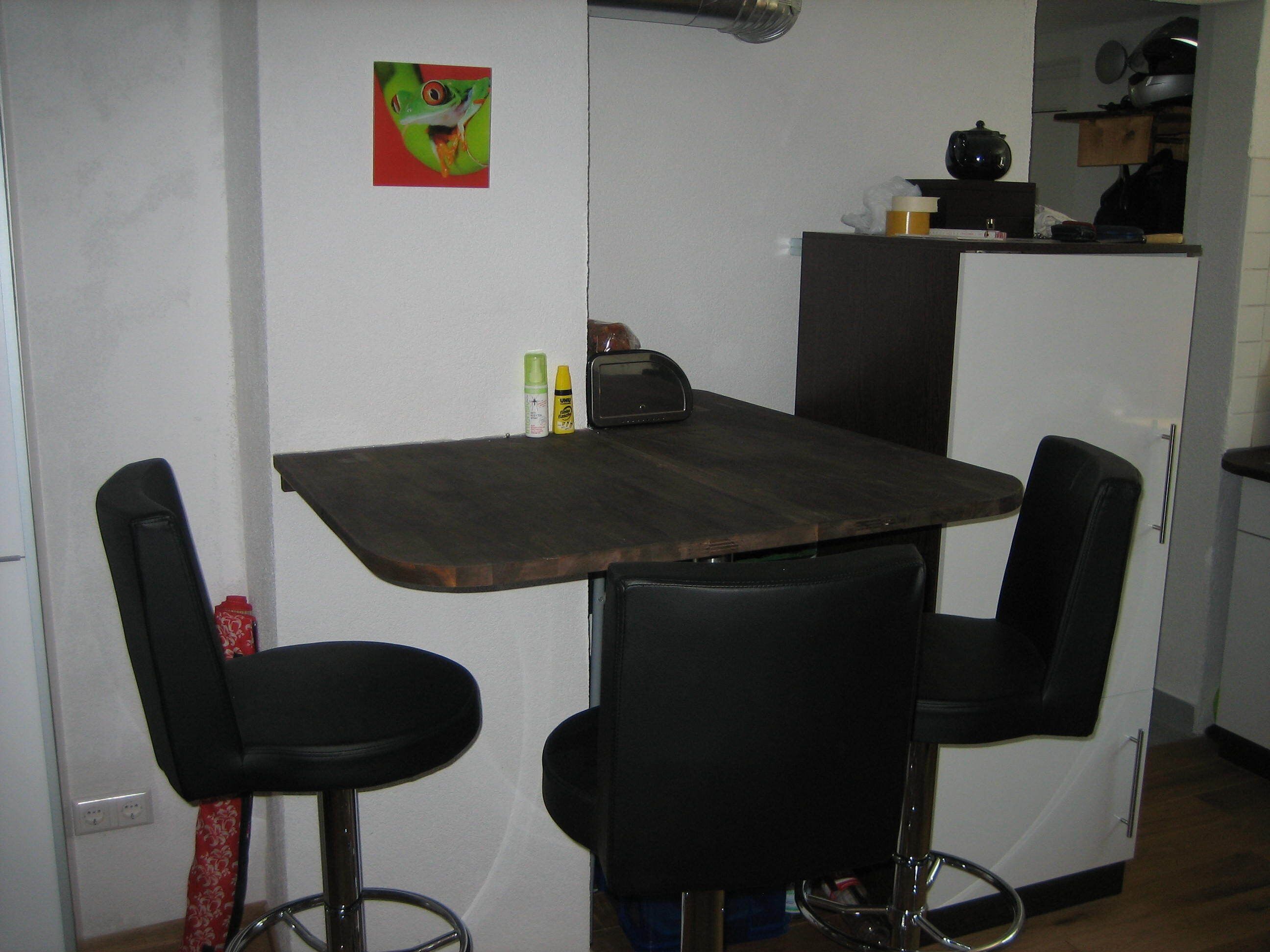 A small kitchen with a wooden table and two black bar stools. There is a small refrigerator and a microwave on the counter.