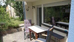 Eine Außenpatio mit Tisch und zwei Stühlen, ein Fenster mit Blick ins Grün und eine kleine Gartenanlage.