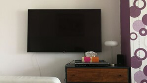 Ein Zimmer mit einem an der Wand montierten Flachbildfernseher, einem Nachttisch aus Holz mit einem Laptop und einem Lautsprecher sowie einem weißen Stuhl.