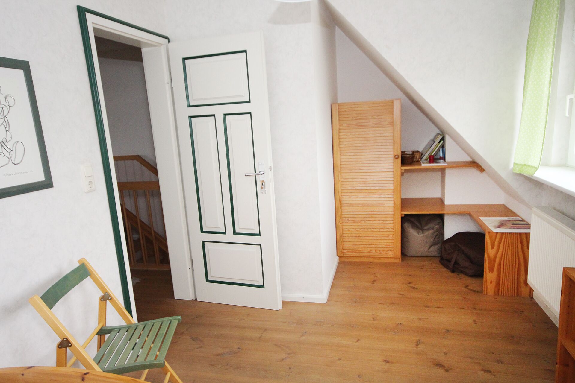 Una habitación con piso de madera, una silla verde, una puerta blanca con detalles en verde, un estante de madera y una ventana con cortina verde.