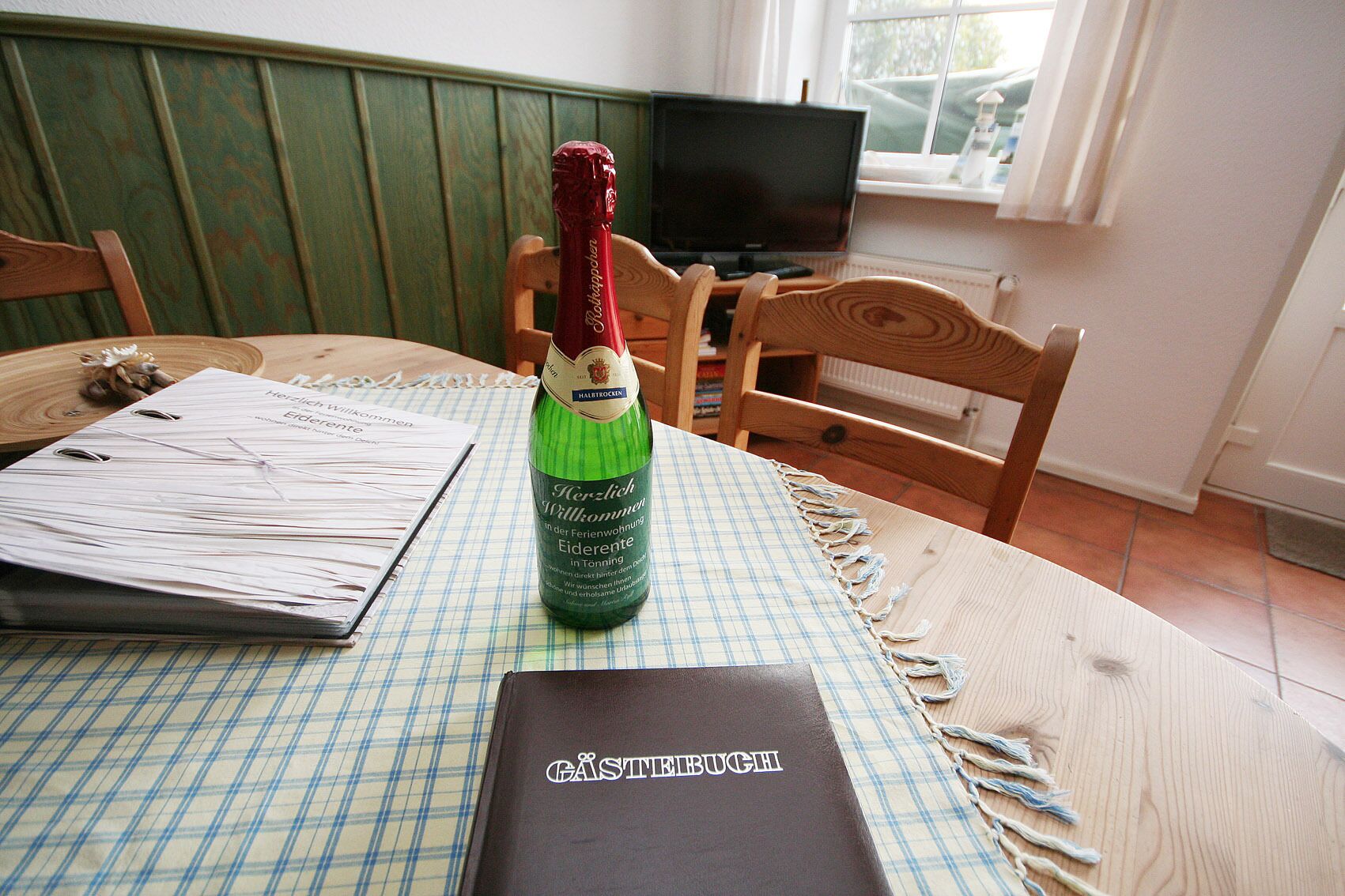 Une table avec une bouteille verte, un classeur noir et une chaise en bois, dans une pièce dont le mur est vert et qui possède une fenêtre.