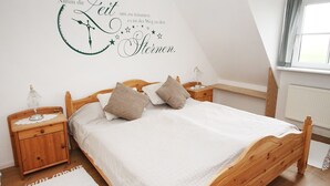 A bedroom with a wooden bed, two bedside tables, a wall clock, and a window with curtains. - Komf. Apartment, 5 pers., 3 bedrooms, 2 bathrooms, near Sankt Peter-O. , right on the dike (Tönning)