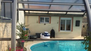 Una piscina cubierta al aire libre con cerramiento de vidrio, rodeada de plantas en macetas y una zona de barbacoa.