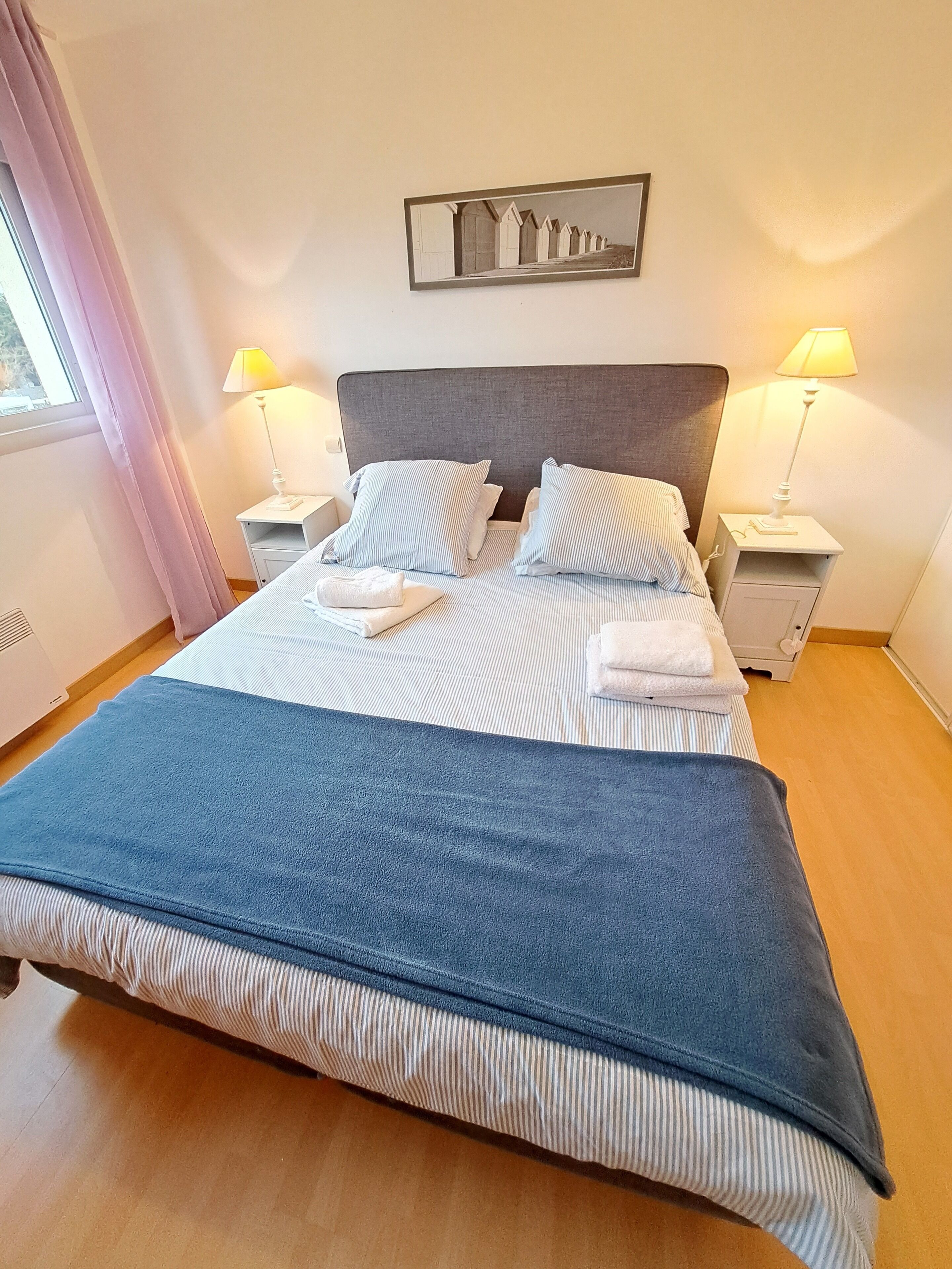 Una cama bien hecha con ropa de cama a rayas y una manta azul, dos lámparas de noche y un cuadro enmarcado sobre la cama.