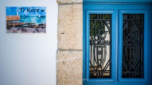 Uma porta azul com detalhada ferragem e uma pintura de paisagem costeira com um farol e o texto "Ti Kere".