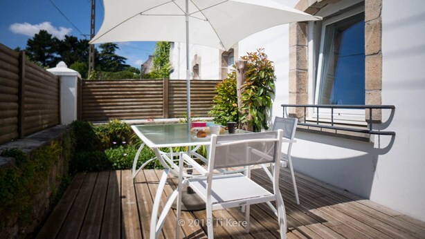 Um pátio com mesa e cadeiras, um guarda-sol branco e um pequeno vaso com planta.