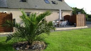 Une maison avec un toit en pente, une terrasse avec une table et des chaises, et un petit jardin agrémenté d’un palmier.