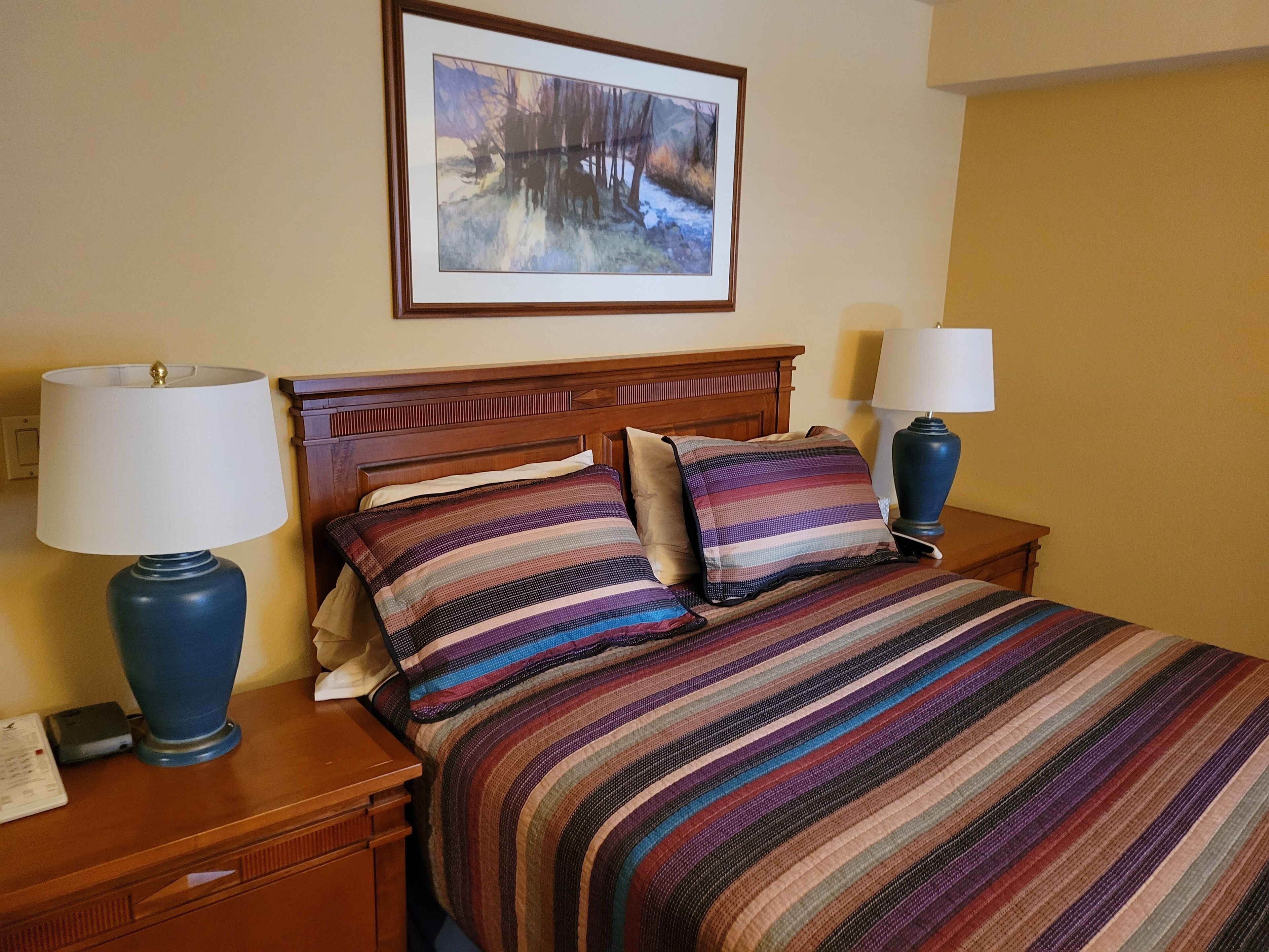 Een hotelkamer met een houten hoofdbord, een bed met gestreept beddengoed, twee bedlampen en een ingelijst schilderij aan de muur.