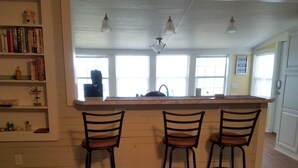 Une cuisine avec un îlot central en bois, trois tabourets de bar et une bibliothèque contenant des livres et des objets décoratifs.