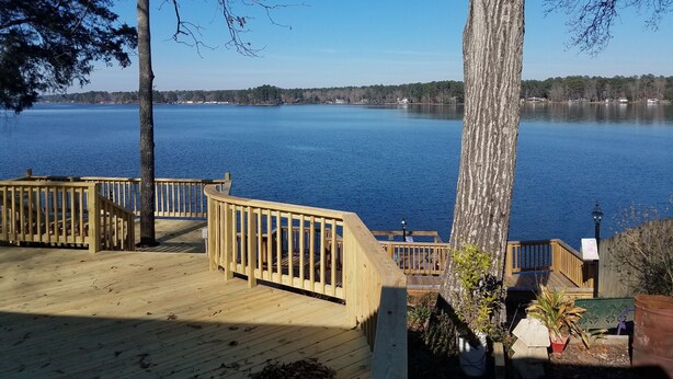 Une terrasse en bois dotée d’une balustrade, qui offre une vue sur un lac paisible, entouré d’arbres et bordé par une rive éloignée.
