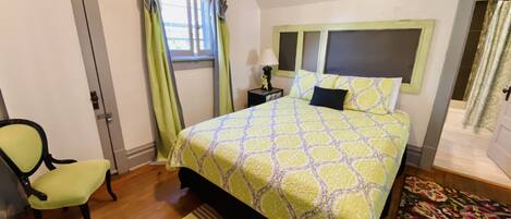 Un dormitorio con cama, una silla, una mesita de noche, una ventana con cortinas y una puerta.