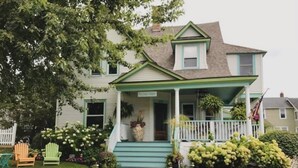 Une maison à deux étages avec un porche à l’avant, des marches menant à l’entrée et un petit jardin agrémenté de plantes et d’un sentier.