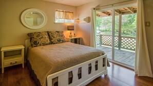 A bedroom with a bed, a nightstand, a mirror, a window with curtains, and a view of a wooden deck.