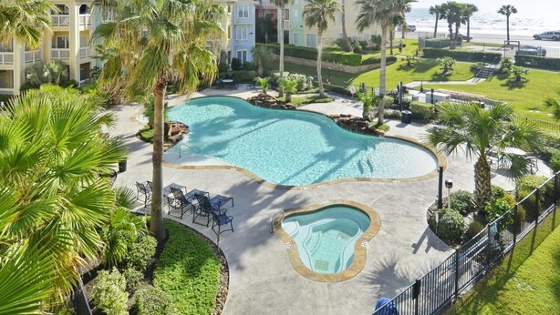 A resort with a large swimming pool, palm trees, and a view of the ocean. - Mermaid Lookout Beachfront Condo (Galveston)