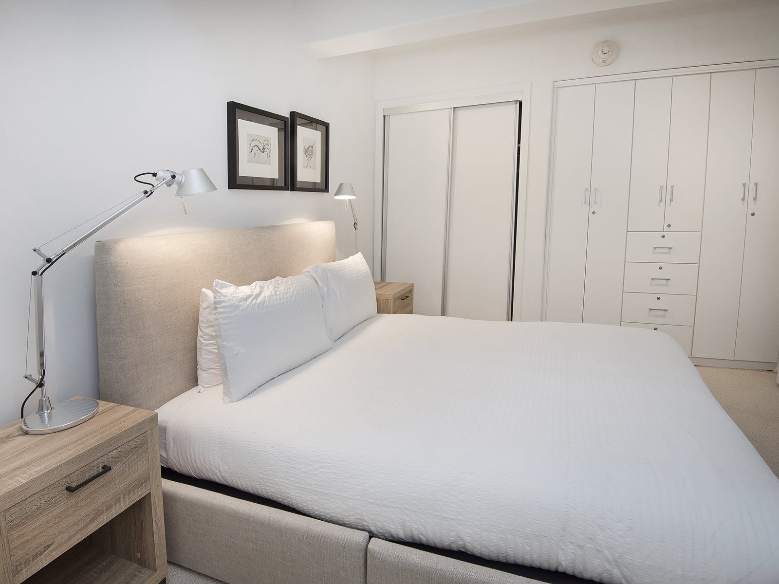 Quarto com cama, mesa de cabeceira, luminária, guarda-roupa e duas imagens emolduradas na parede.