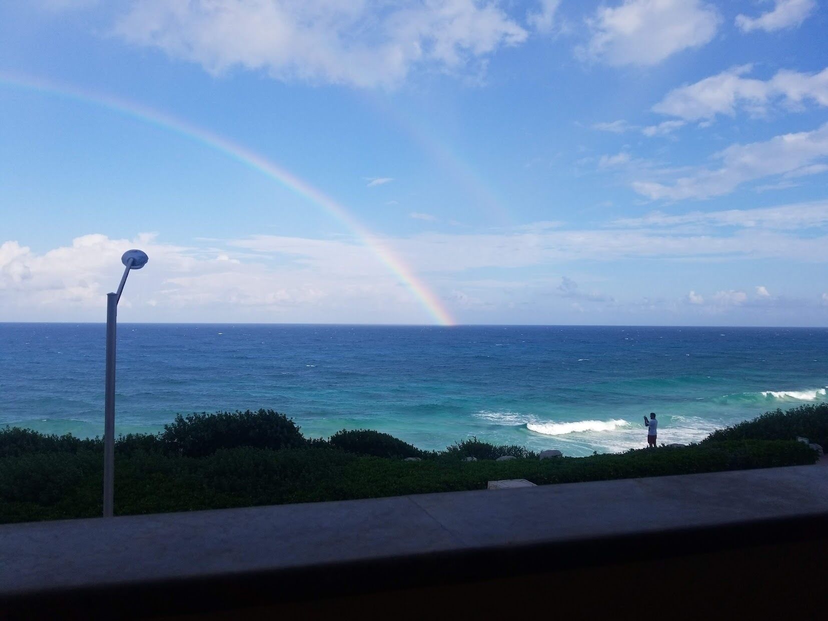 A view of the ocean with a rainbow and a person taking a photo.