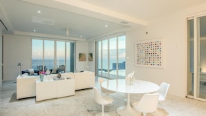 Een moderne woonkamer met een witte bank, een ronde eettafel met witte stoelen, en grote ramen met uitzicht op de oceaan.