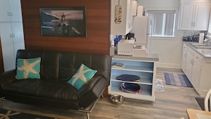 A living room with a black leather sofa, teal and gold cushions, a framed picture on the wall, and a kitchen area with white cabinets, a refrigerator, and a sink.