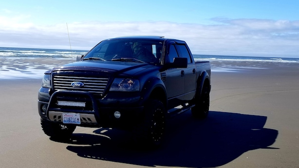 A black Ford F-150 truck parked on a sandy beach with ocean waves in the background.