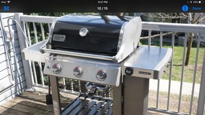Una parrilla Weber Genesis II portátil sobre una terraza de madera con una barandilla al fondo.