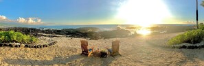 Una spiaggia con due sedie di legno e un tavolino con bevande, circondata da rocce e vegetazione.