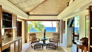 A spacious room with a large window offering a view of the ocean and palm trees. There are two leather chairs and a small round table with a statue. The room has wooden cabinets and a ceiling fan. - Exotic Kona Bay Hawaii Beachfront Tropical Paradise Stunning Home. (Kailua-Kona)